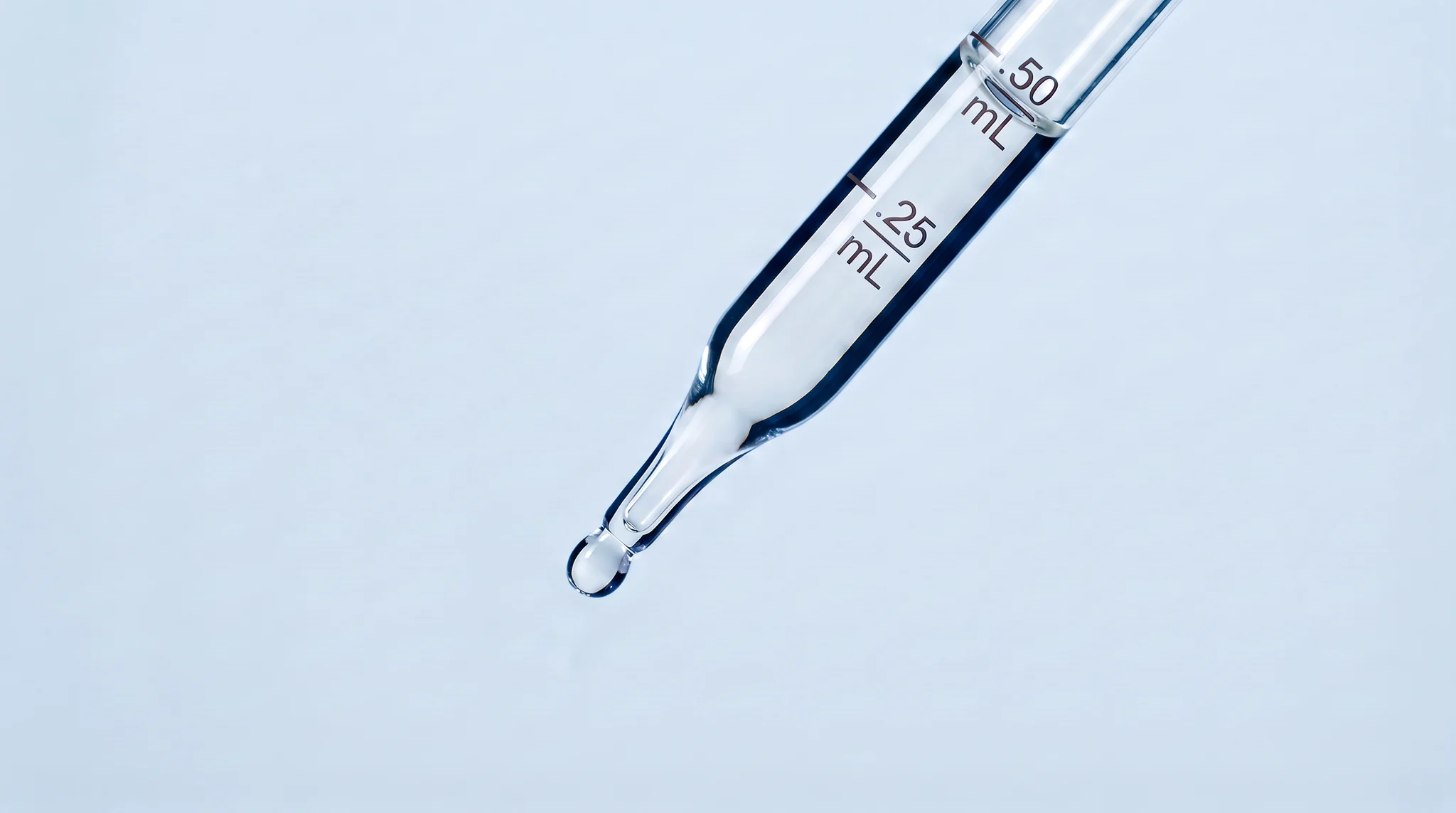 A glass pipette with a drop forming at its tip, against a pale blue background